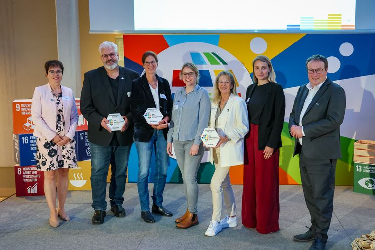 Gruppe von sechs Personen bei einer Veranstaltung mit Preisen und buntem Hintergrund.